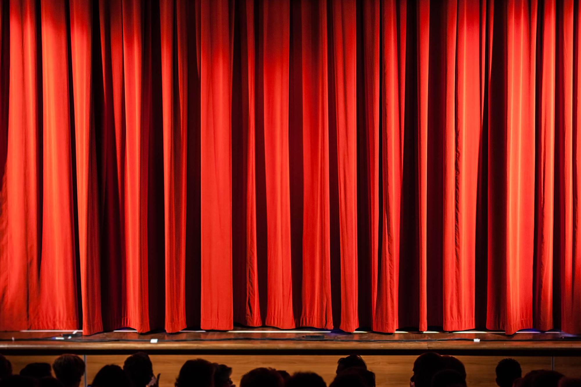 Theatre stage with rippling curtains, overhead lights, and silhouetted audience in the foreground