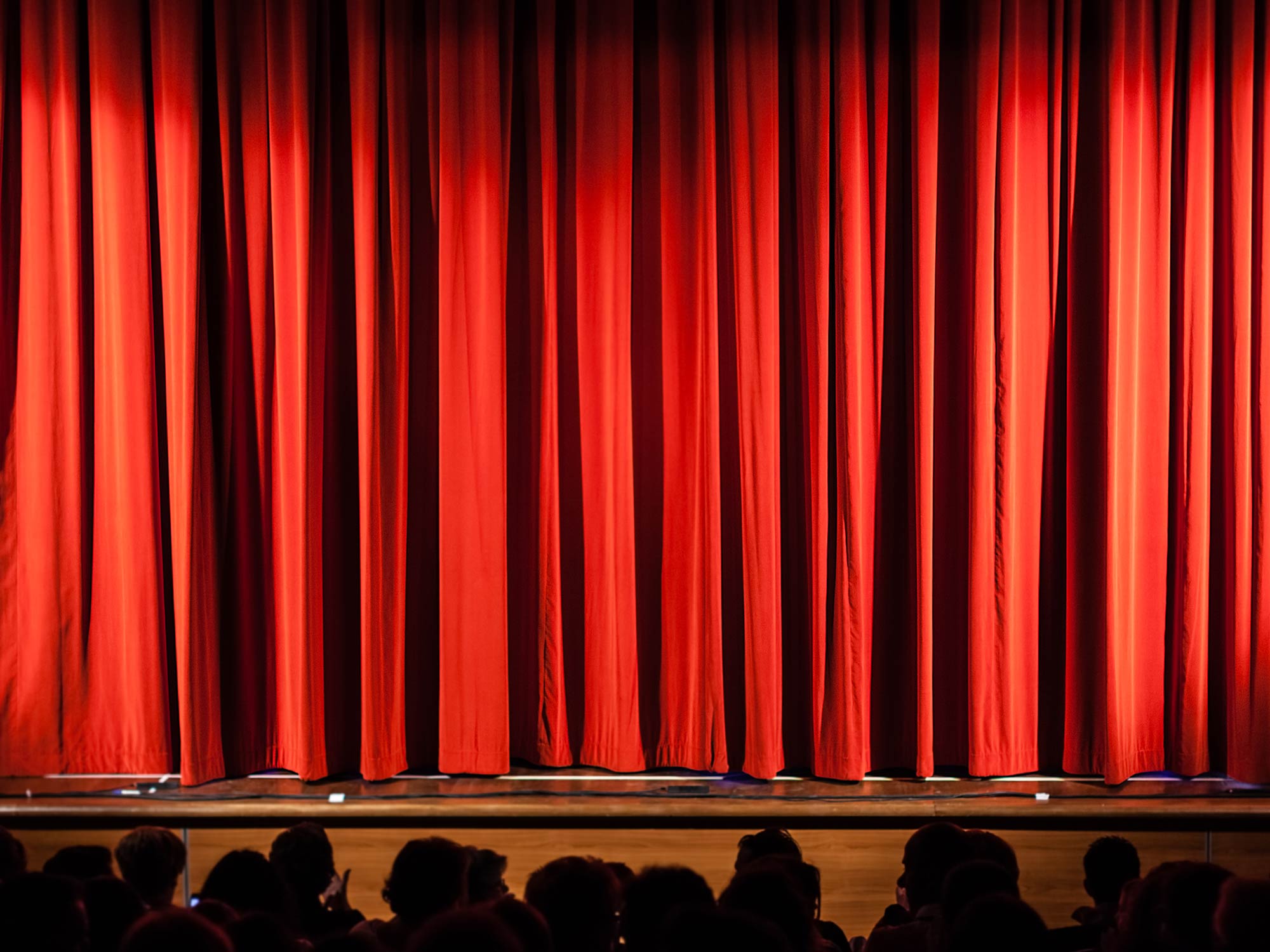 Theatre stage with rippling curtains, overhead lights, and silhouetted audience in the foreground