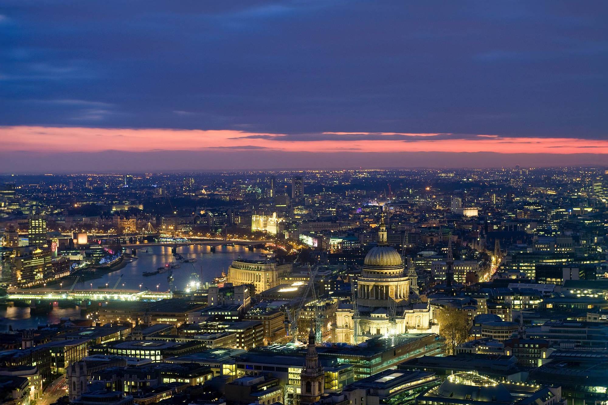 Aerial view of the expansive London skyline at dusk, with city lights illuminating the streets beneath a sky with scattered clouds