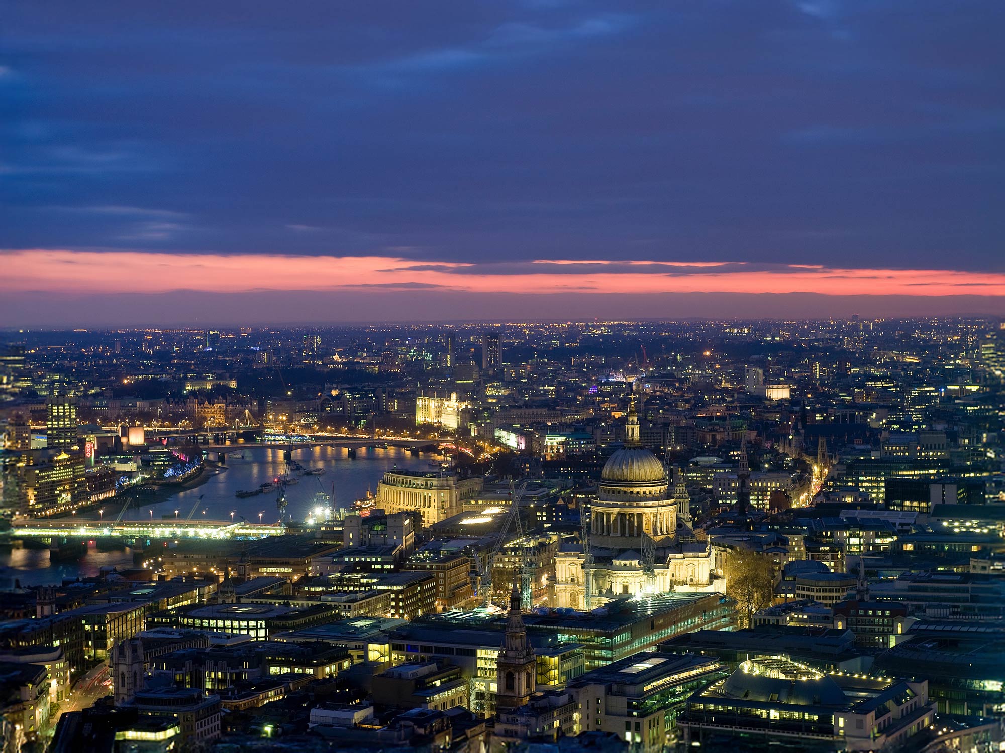 Aerial view of the expansive London skyline at dusk, with city lights illuminating the streets beneath a sky with scattered clouds